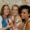 Three women holding colorful striped bottles against a stone wall.