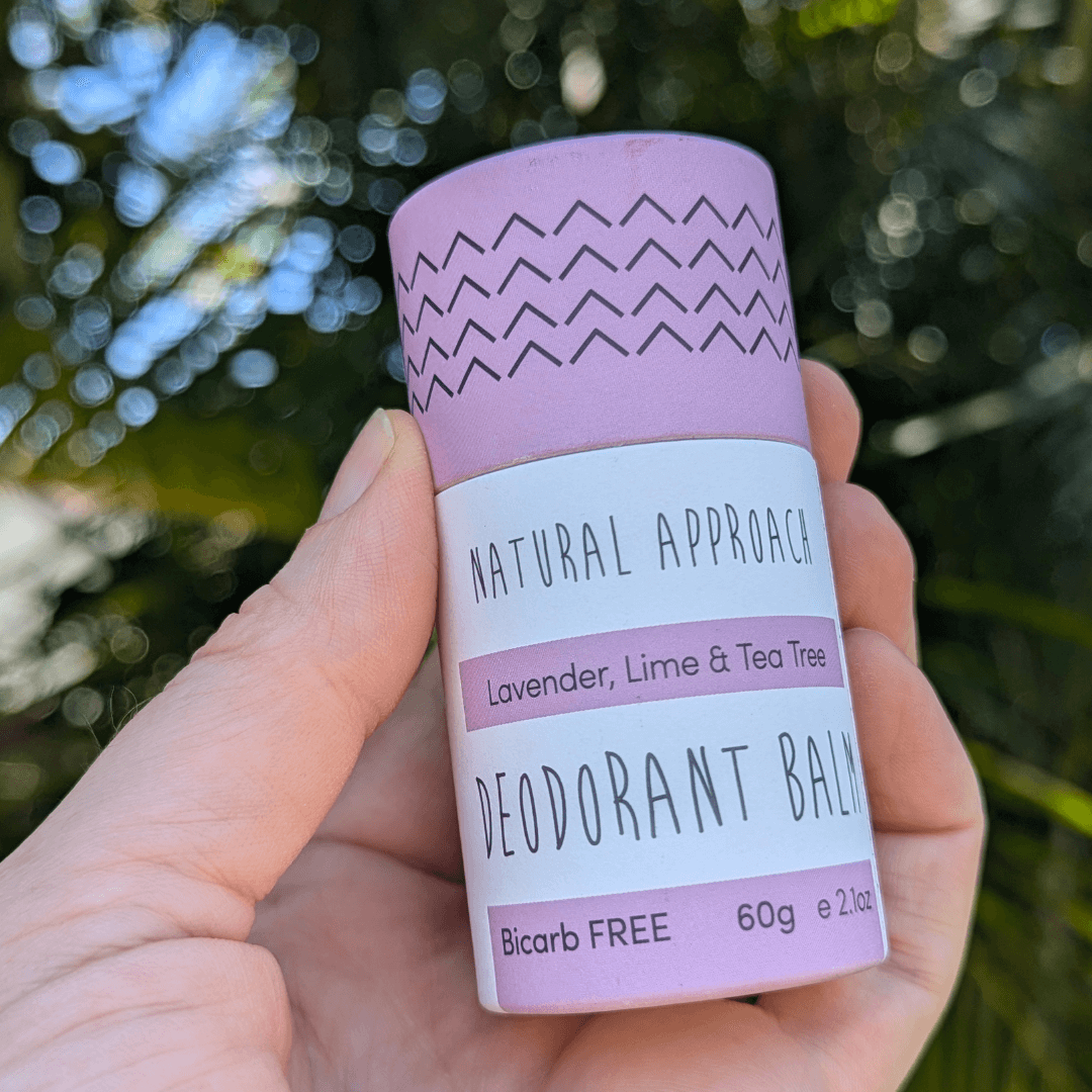 Hand holding a Natural Approach Lavender, Lime & Tea Tree deodorant balm with a blurred natural background of trees
